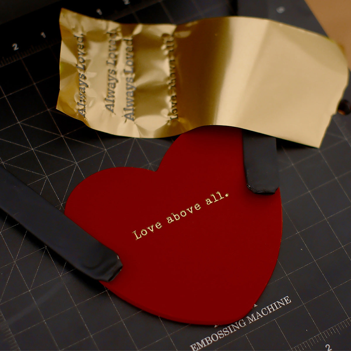 Red heart-shaped keychain with 'Love above all' embossed in gold foil, displayed alongside an embossing machine and gold foil material.