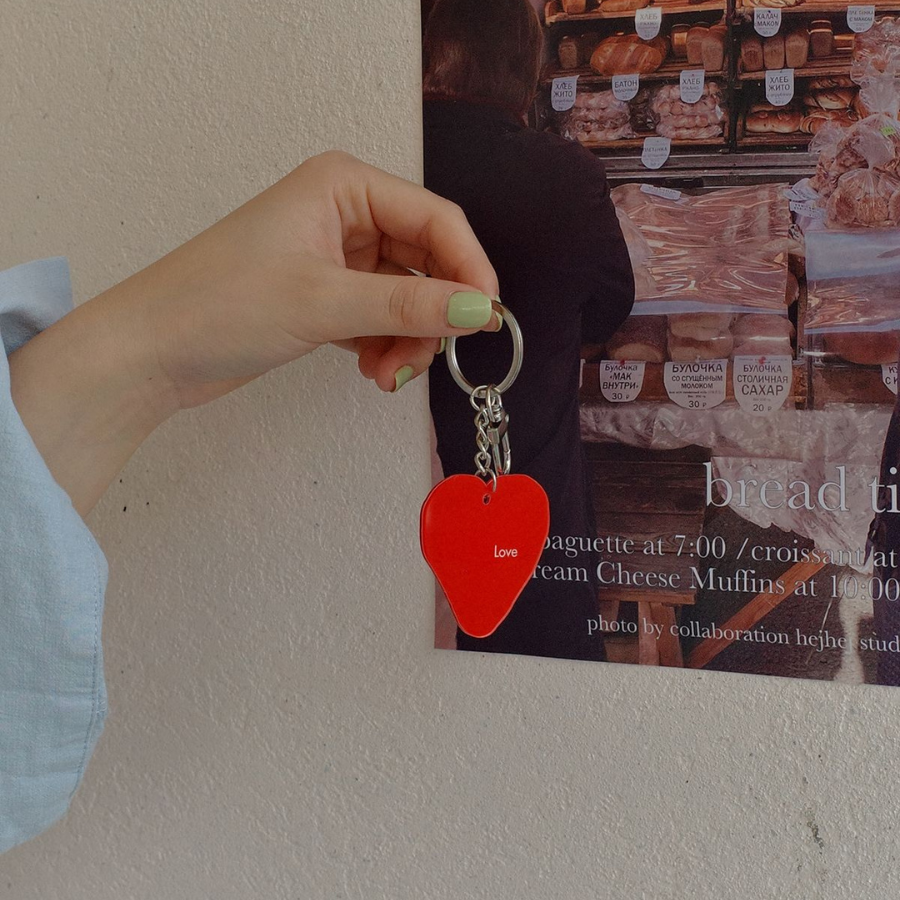 Hand holding a red heart-shaped keychain with "Love" inscribed, against a bakery-themed poster, symbolizing affection and faith.