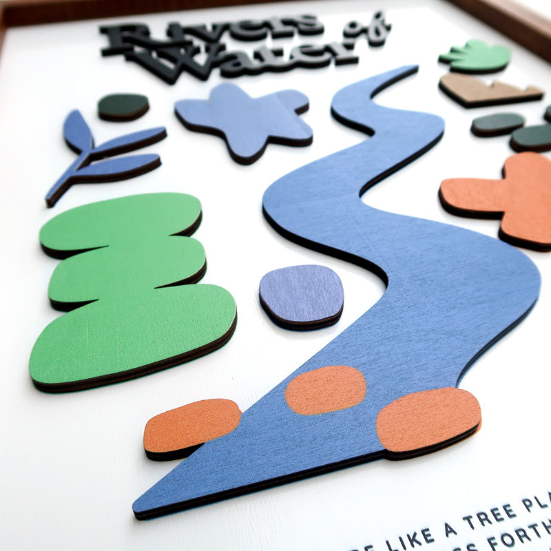 Close-up of a handcrafted wooden scripture poster featuring "Rivers of Water" from Psalm 1:3. The design includes layered elements like a blue river, colorful shapes, and natural motifs, framed in wood.
