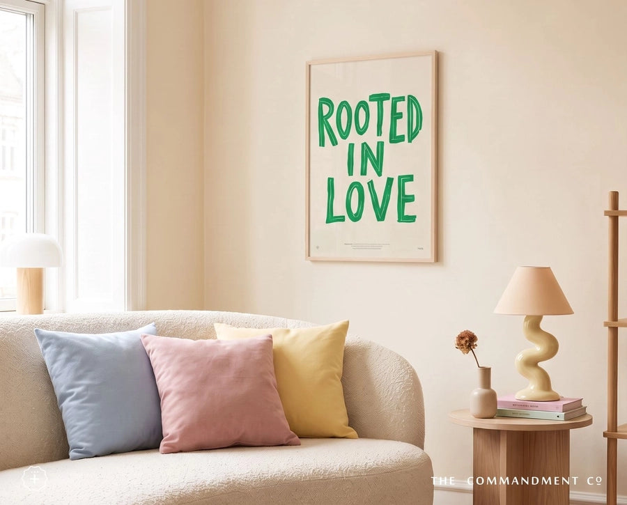 Rooted In Love Christian poster displayed above a cream sofa in a warm living room with pastel cushions and a table lamp