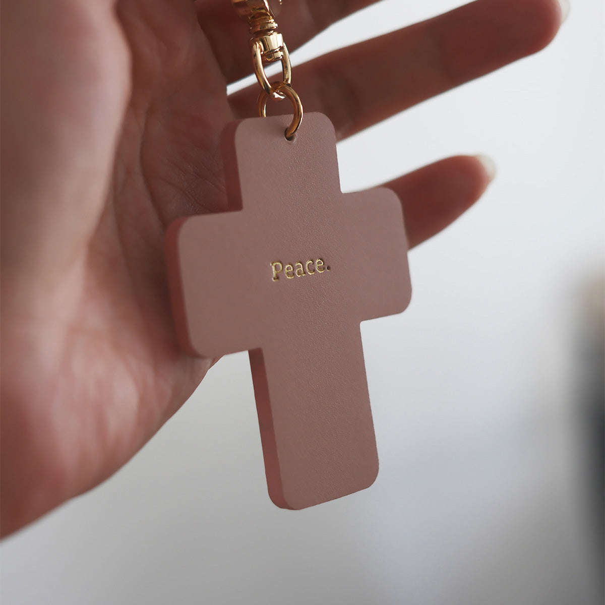 Rose pink cross-shaped keychain with 'Peace' engraved, held in a hand, symbolizing faith and serenity.