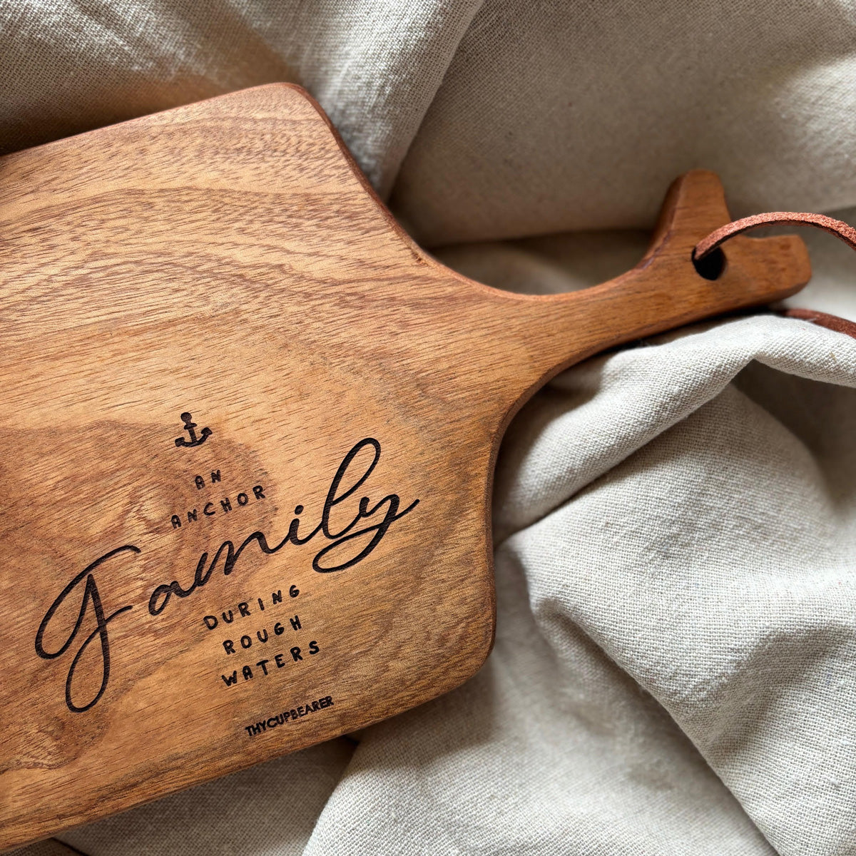 A wooden cheese board featuring the engraved phrase "Family, an anchor during rough waters" with an anchor icon. The board highlights its rich wood grain and leather strap handle, placed against a soft beige fabric. Perfect for rustic kitchen decor or as a meaningful family gift.