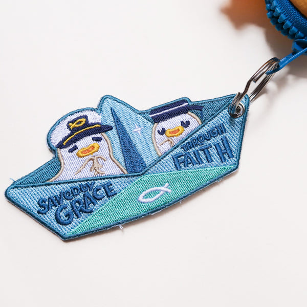 Saved by Grace Through Faith embroidered Christian keychain shaped like a paper boat with cute sailor characters attached to a bag zipper.