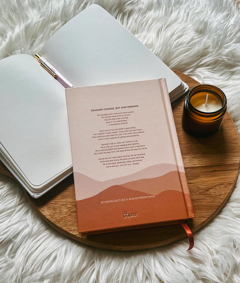 Hardcover journal with printed message on back cover by Project J placed on wooden tray beside open notebook and candle.