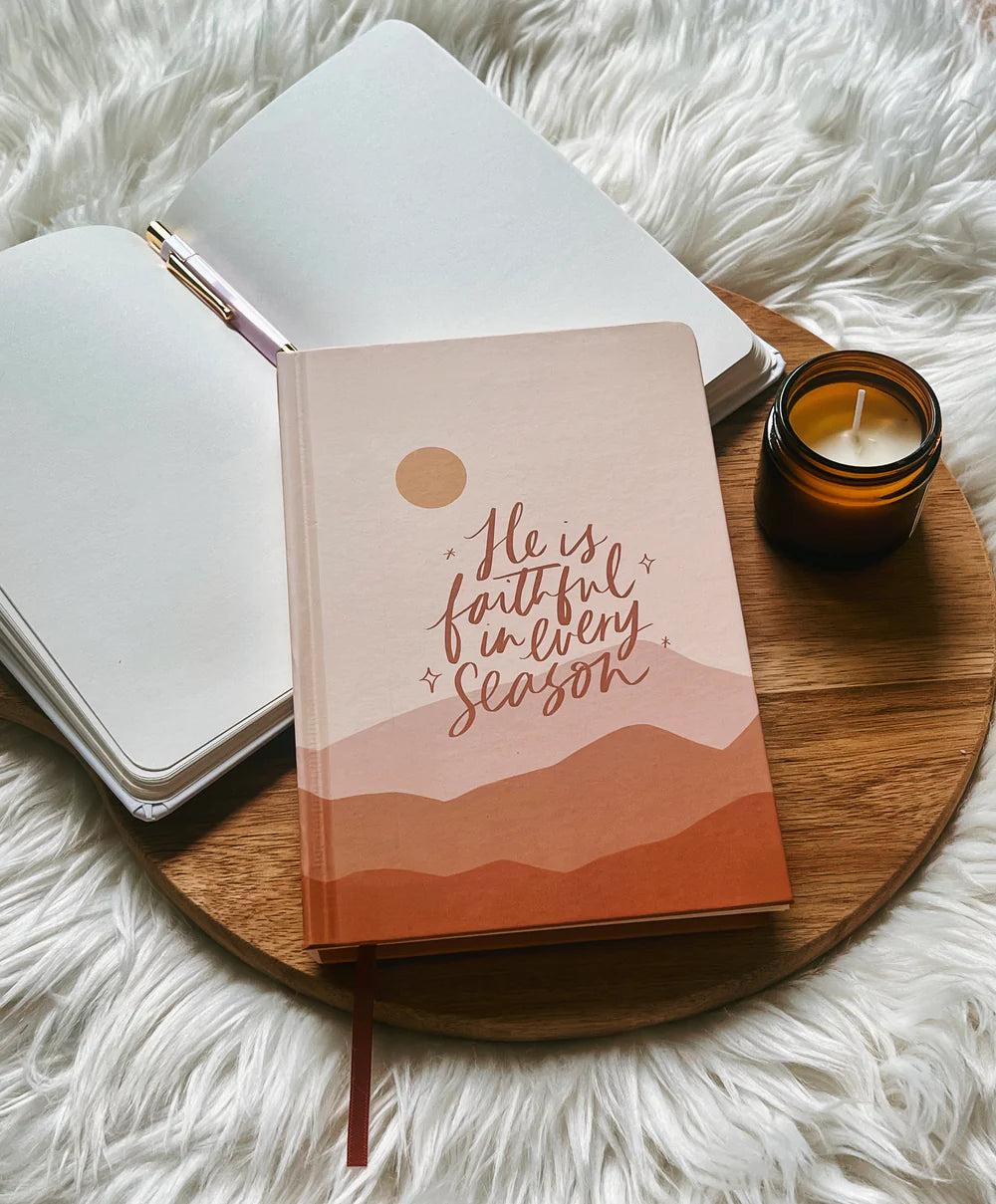 Hardcover journal with “He is faithful in every season” text design by Project J placed on wooden tray beside open notebook and candle.