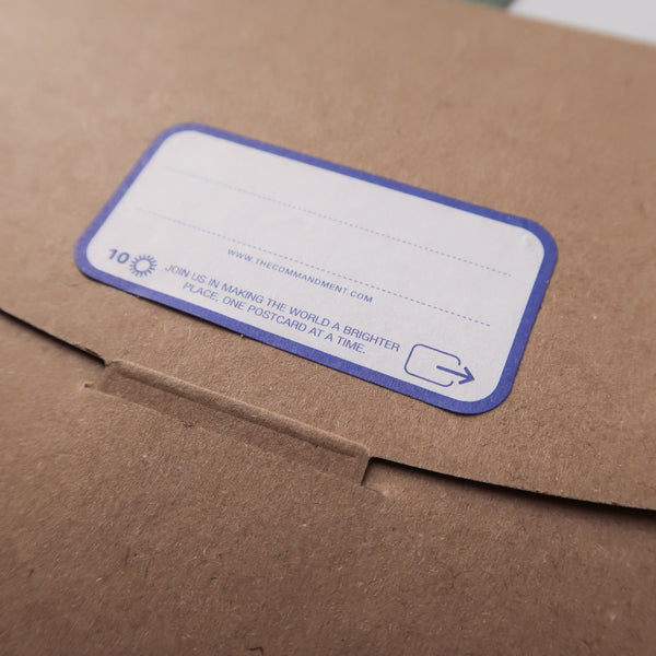 A close-up of a brown envelope with a label reading, "Join us in making the world a brighter place, one postcard at a time," encouraging positivity and connection through postcard sharing.