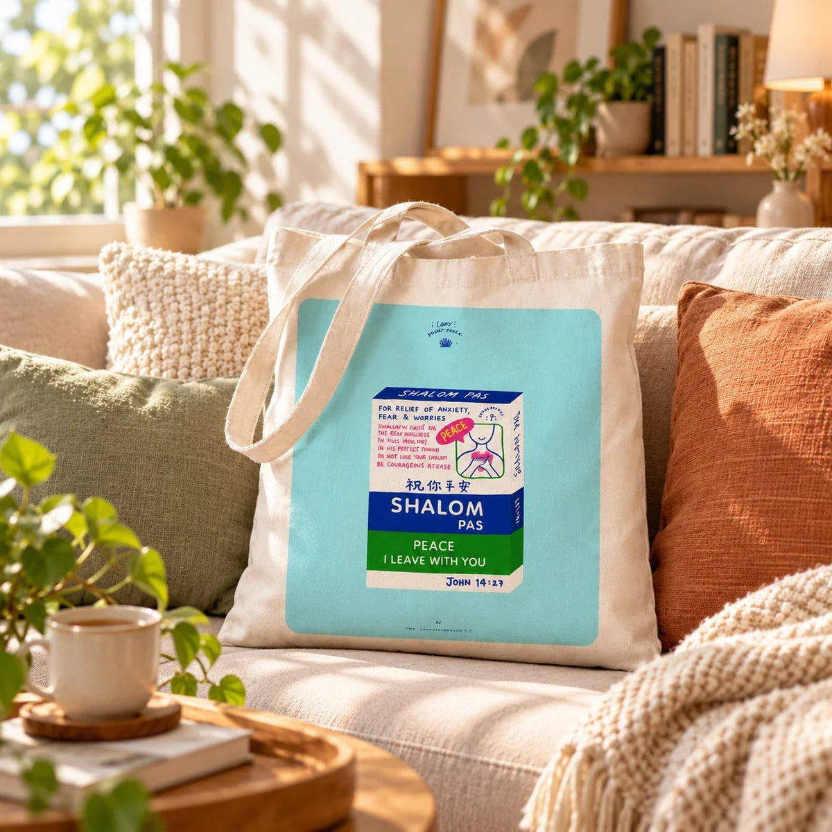 Shalom Pas Christian tote bag on a cosy beige sofa in a sunlit living room, with canvas handles draped down.