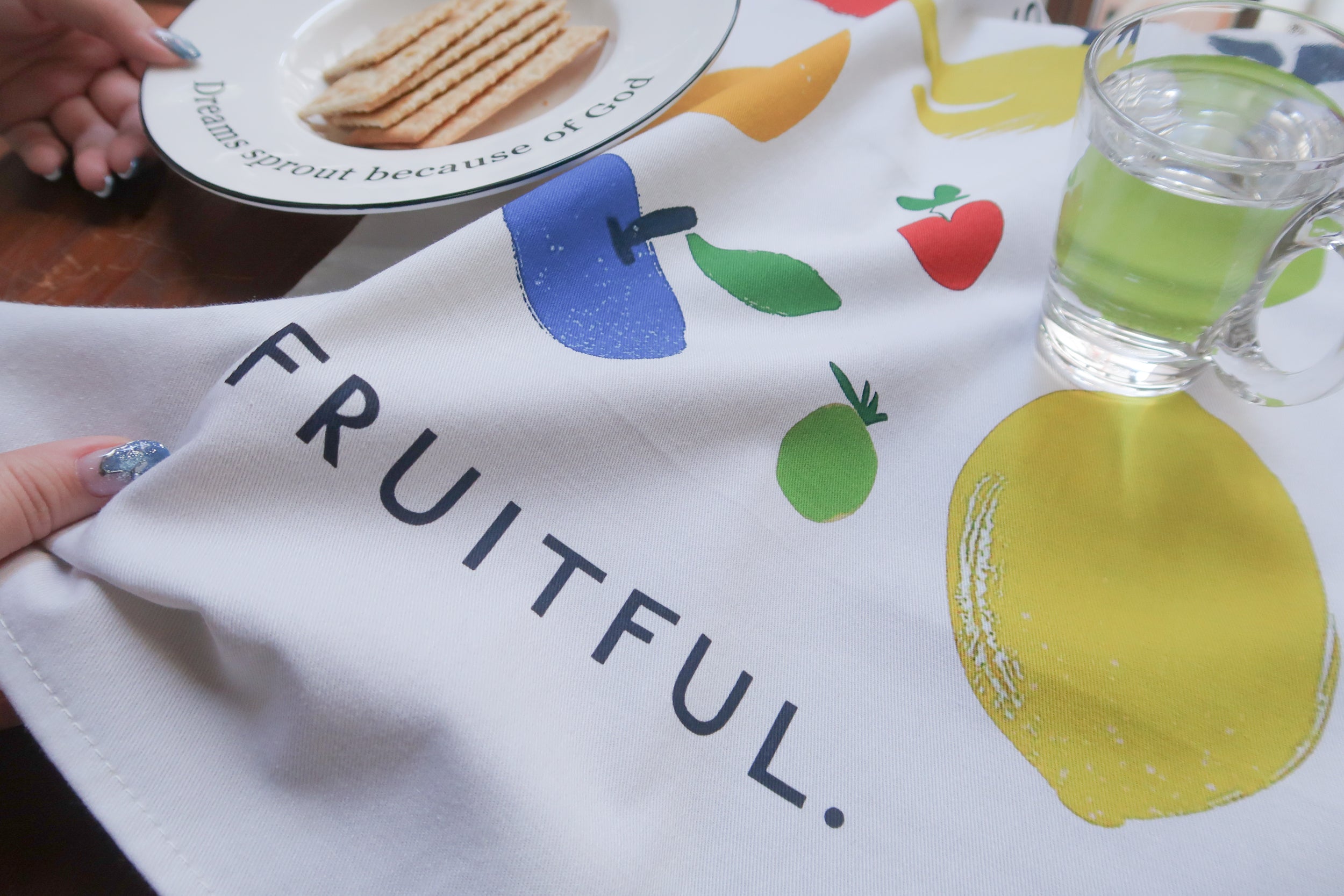 Stay Fruitful tea towel featuring a colorful fruit print (red apple, yellow lemon, green pear, blue plum, and small green fruit) on white fabric; faith-inspired kitchen decor by The Commandment Co.