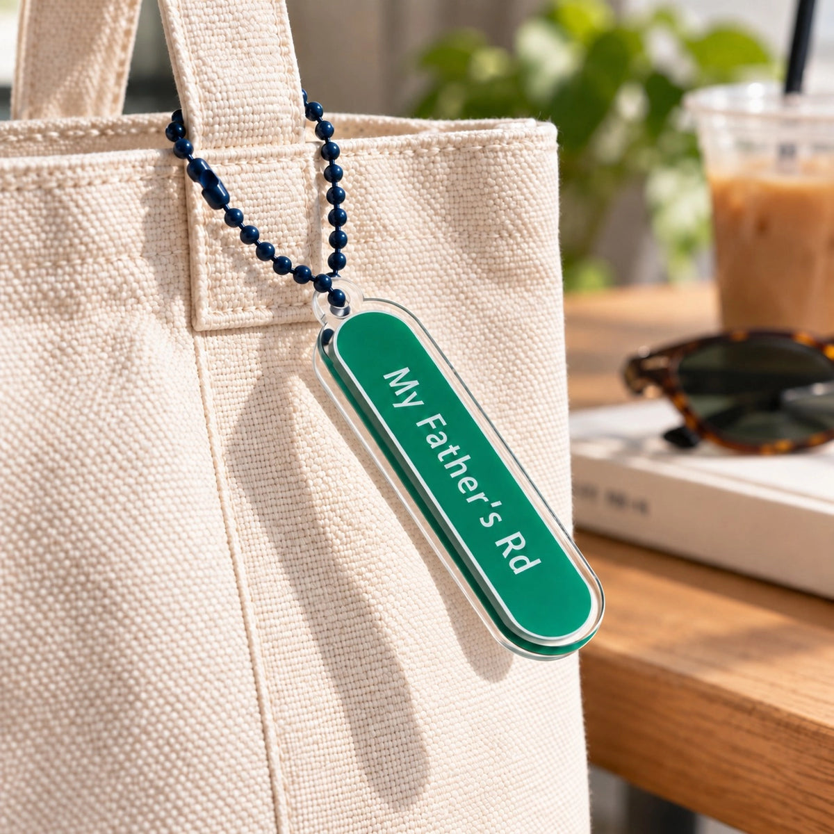 My Father’s Road green acrylic street-sign keychain hanging from a cream canvas tote bag in a café setting.