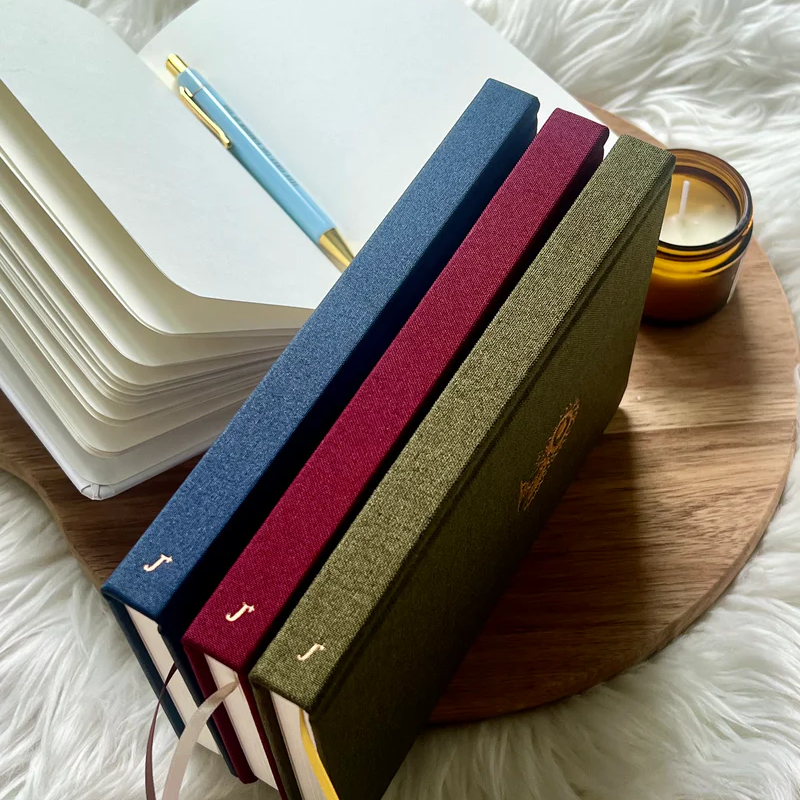 Three fabric hardcover journals by Project J in blue, red, and green displayed beside an open notebook and candle on a wooden tray.
