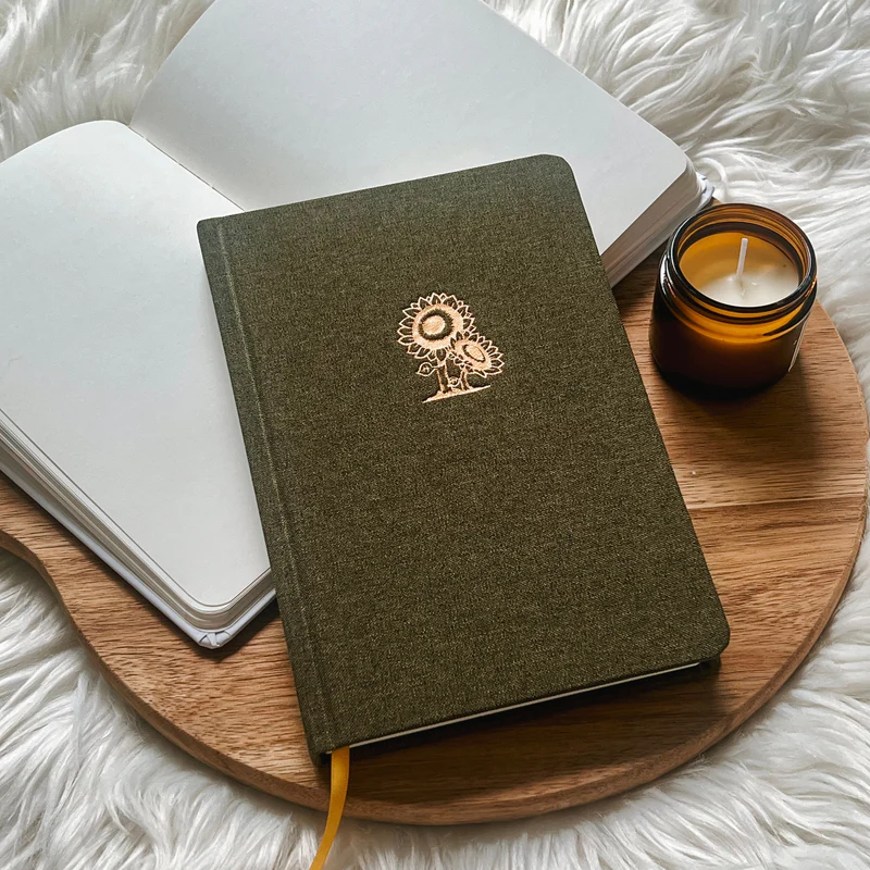 Fabric hardcover journal with gold sunflower design placed on wooden tray with candle and open notebook.