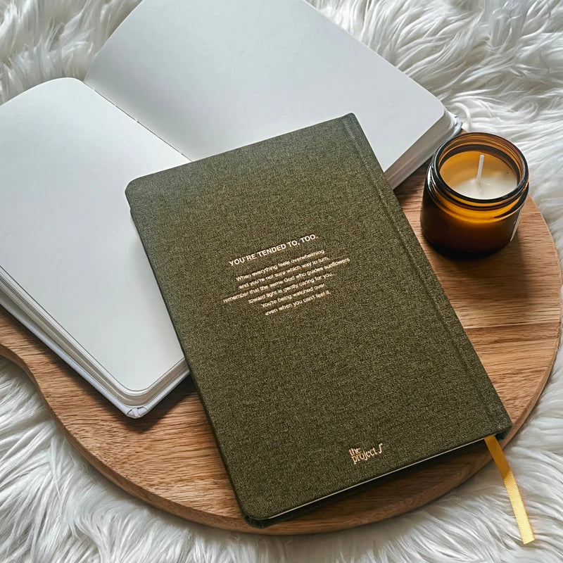 Olive green fabric journal with gold text on back cover by Project J placed on wooden tray beside open notebook and candle.