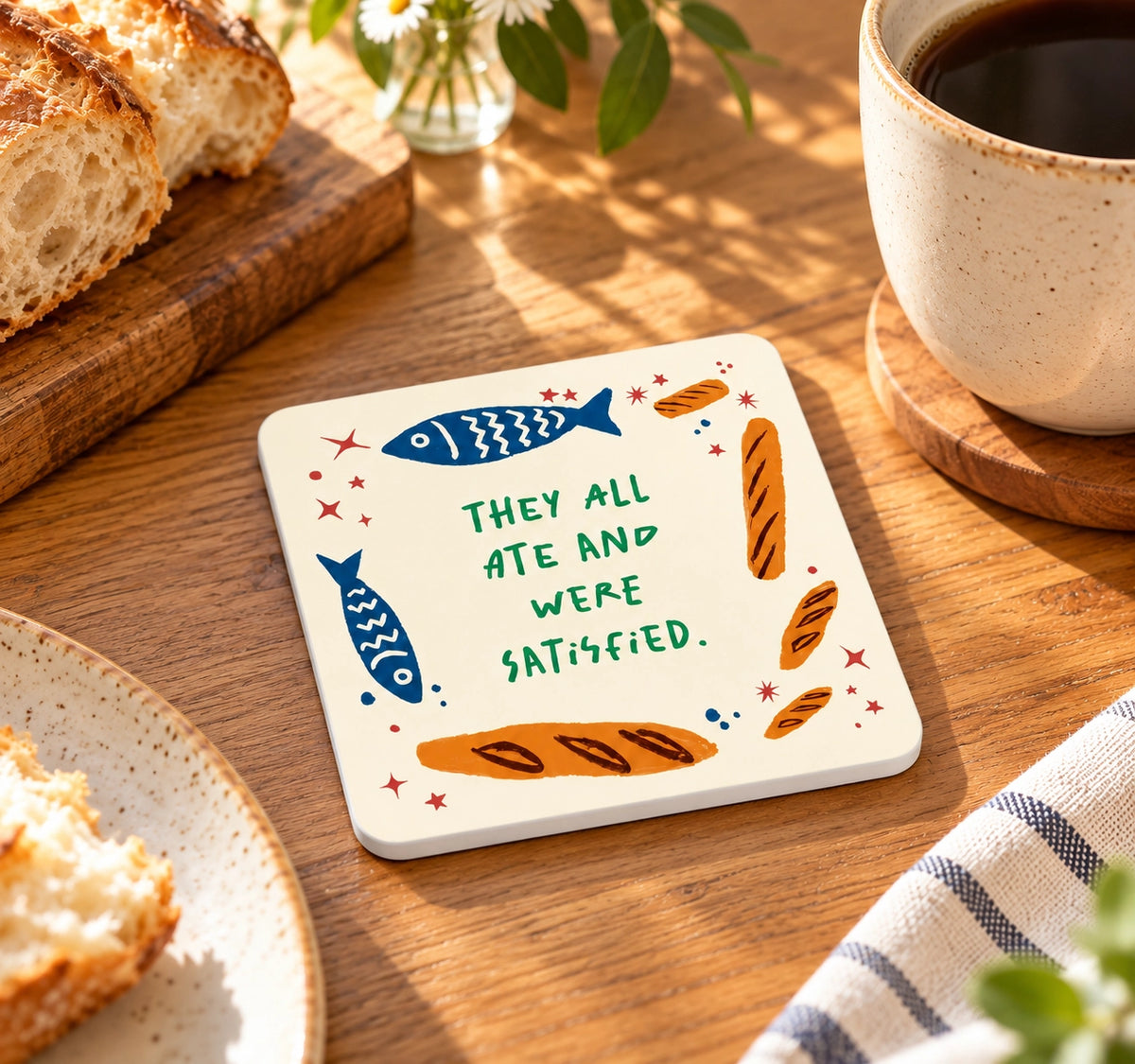 They All Ate And Were Satisfied Christian coaster with fish and loaves illustration beside coffee and bread.
