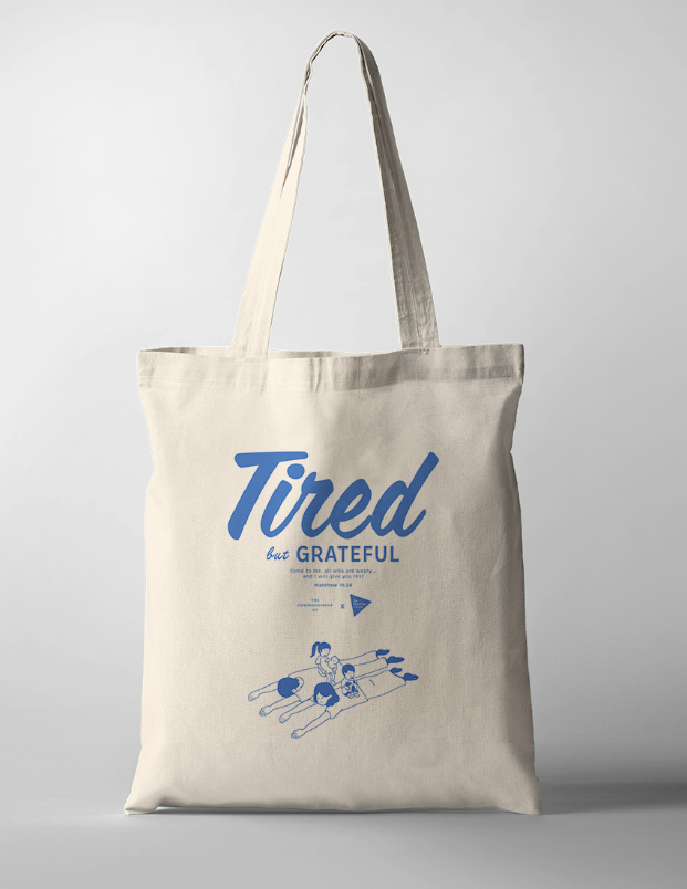 Tote Bag with the phrase "Tired But Grateful" featuring an illustration of parents lying down with two kids on them — a heartfelt design celebrating parenthood and faith.