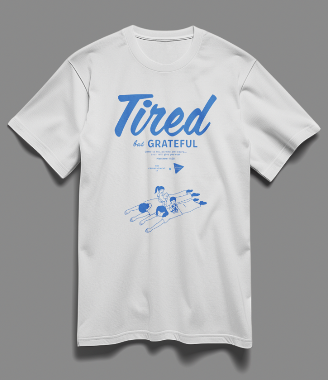 Christian t-shirt with the phrase "Tired But Grateful" featuring an illustration of parents lying down with two kids on them — a heartfelt design celebrating parenthood and faith.