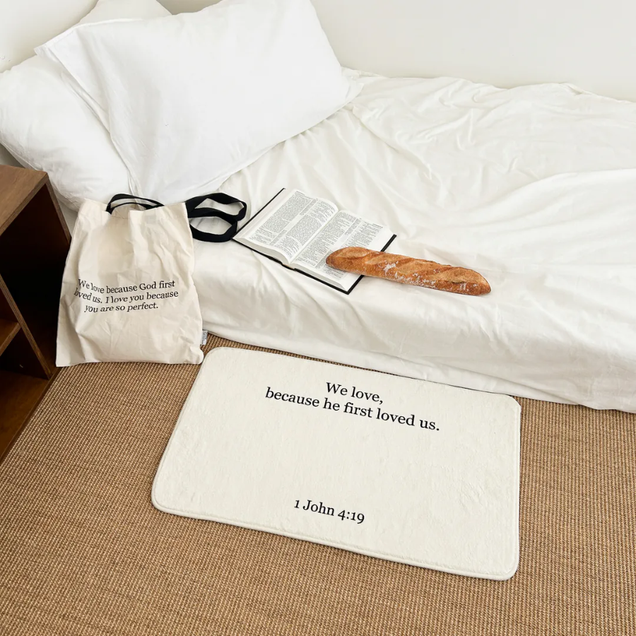 White rug with the words "We love because he first loved us. 1 John 4:19" placed beside a cozy bed, open Bible, and tote bag, inspiring faith and simple happiness as a Christian home decor gift from Jincha God.