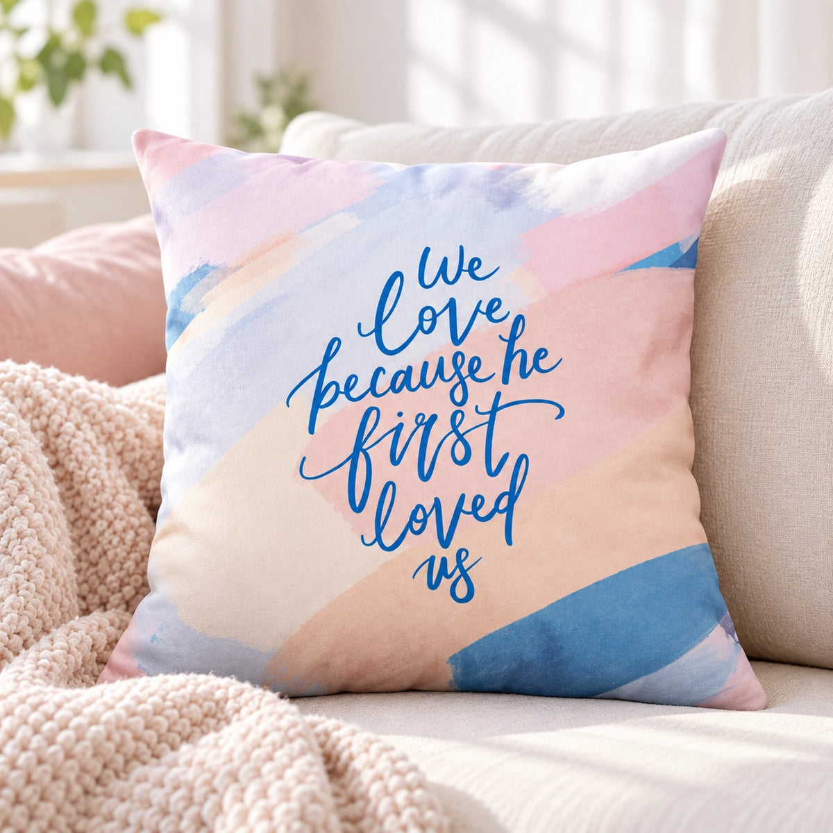 We Love Because He First Loved Us Christian cushion cover styled on a neutral sofa in a bright living room.