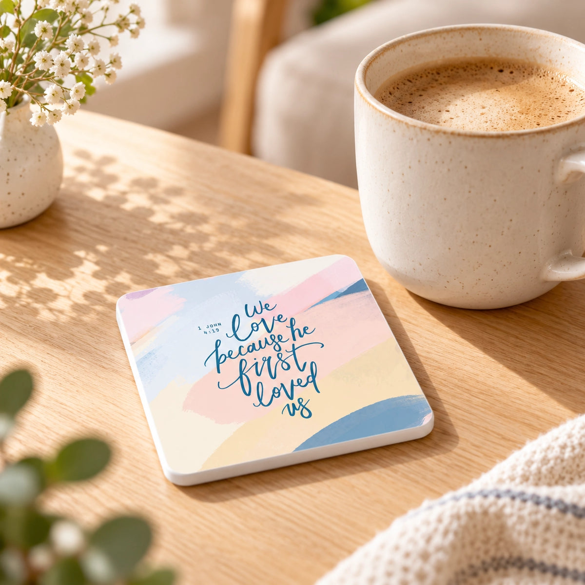 Because He First Loved Us Christian coaster styled beside a ceramic coffee mug on a warm wooden table