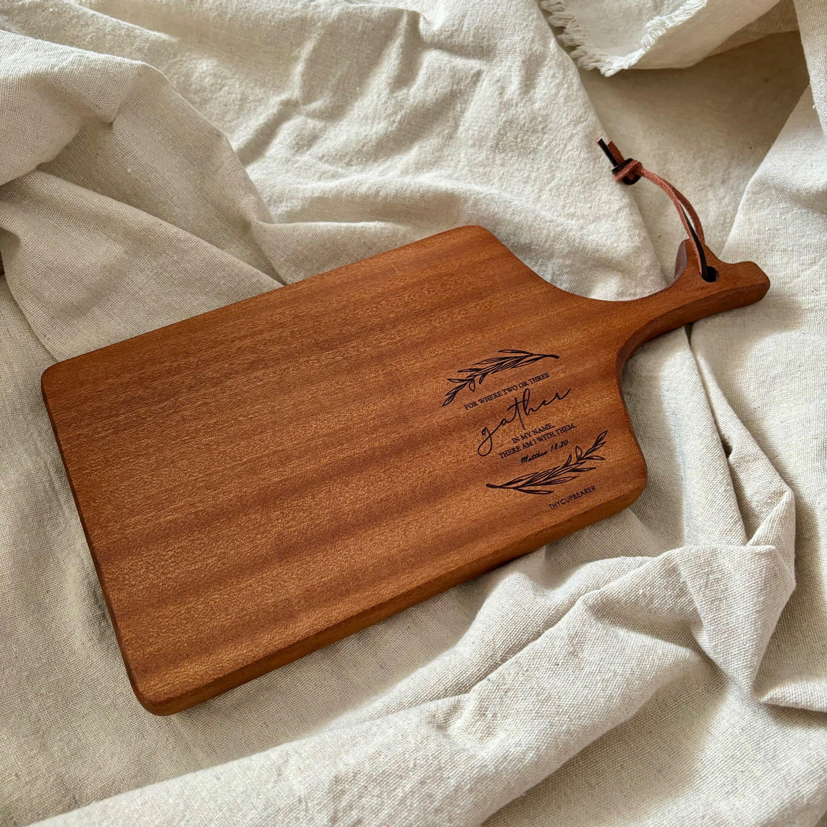 A wooden cheese board engraved with the Bible verse "For where two or three gather in My name, there am I with them" (Matthew 18:20), accented with olive branch illustrations and a leather strap. It rests on a soft beige fabric, highlighting its rustic and elegant design, perfect for Christian home decor or as a meaningful gift.
