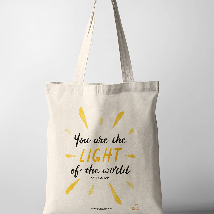 Christian tote bag featuring the Bible verse “You are the light of the world” Matthew 5:14 with yellow sunburst design, collaboration with Chari Designs.