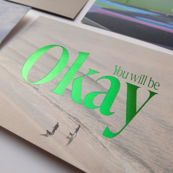 A close-up view of a collection of postcards, with a focus on the "You Will Be Okay" card featuring a serene beach background and bold green text, offering reassurance and encouragement.
