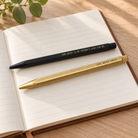 Hexagon Click personalised brass and black ballpoint pens resting on an open lined notebook in warm natural light