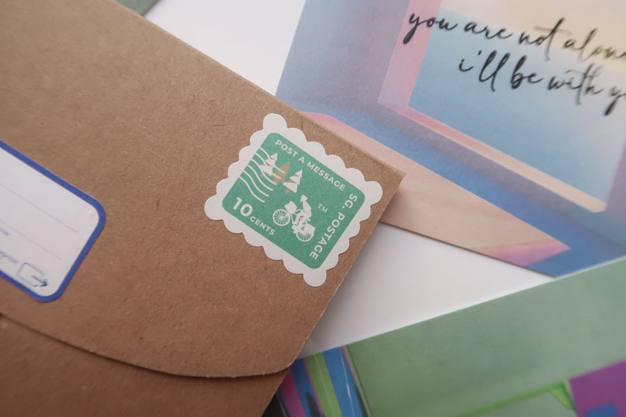 A close-up of an envelope adorned with a 10-cent postage stamp reading "Post a Message," accompanied by a colorful postcard with the encouraging text "You are not alone, I'll be with you.