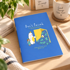Don't Paiseh A6 Christian notebook with blue cover styled on a warm sunlit desk