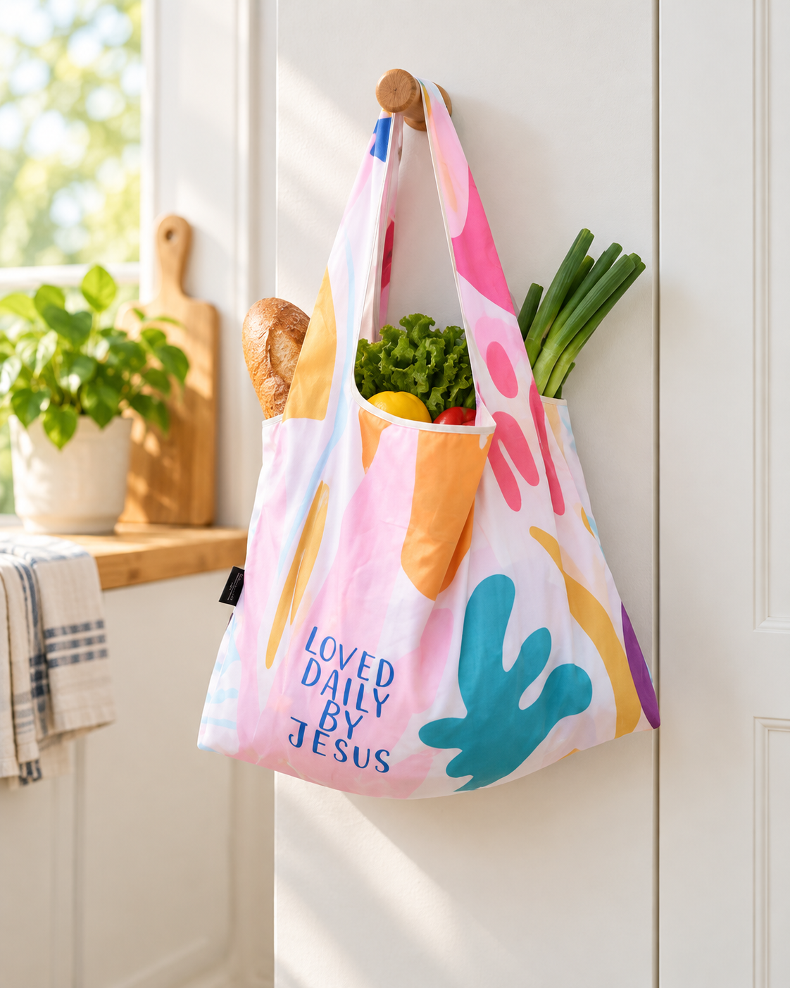 Loved Daily by Jesus reusable Christian shopping bag filled with groceries and hanging in a bright home kitchen