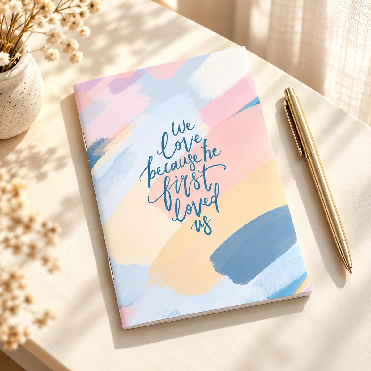 We Love Because He First Loved Us A6 Christian notebook styled on a bright desk with a pen