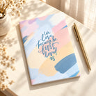 We Love Because He First Loved Us A6 Christian notebook styled on a bright desk with a pen