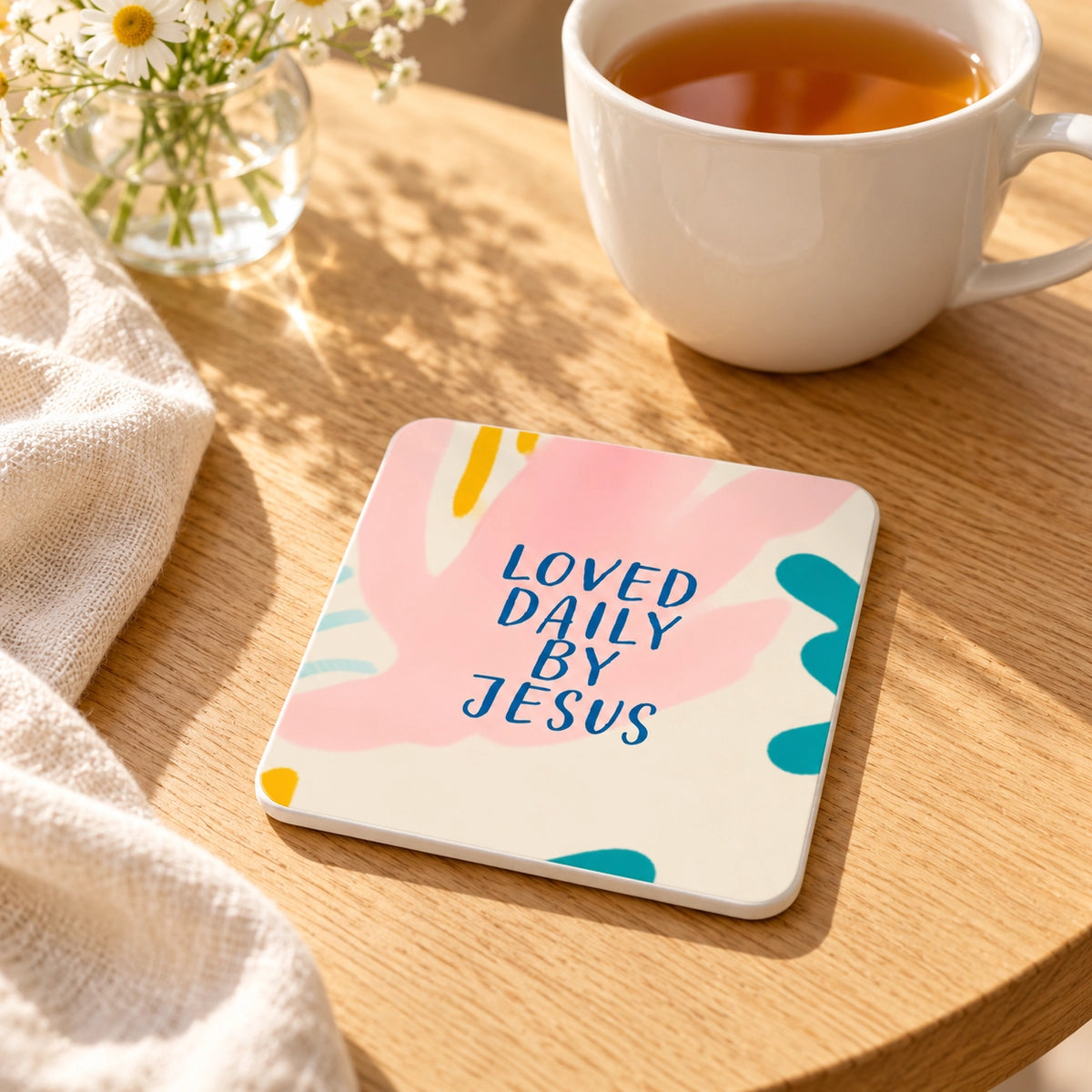 Loved Daily by Jesus Christian coaster styled on a warm wooden table beside a cup