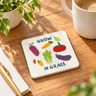 Grow in Grace illustrated vegetable coaster with white side edges on a wooden table beside a coffee mug.