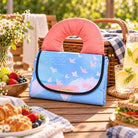 The Lord Will Watch Over Your Coming and Going picnic mat folded beside a wicker basket on an outdoor picnic table