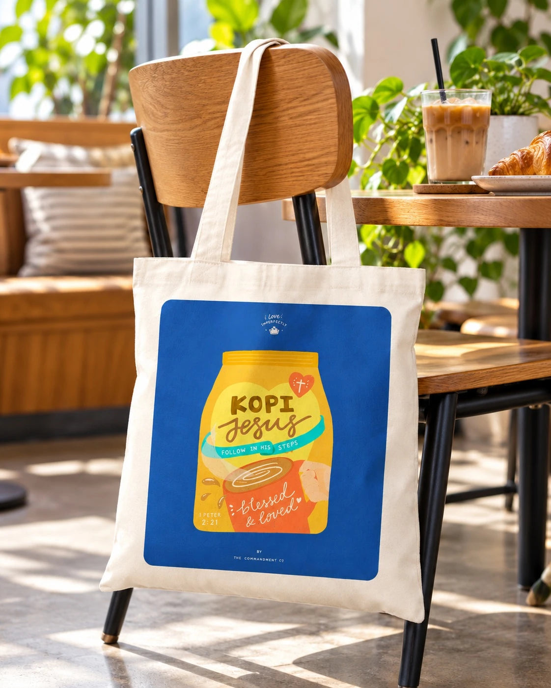 Kopi Jesus Coffee tote bag hanging from a café chair beside an iced coffee