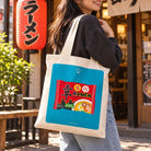 Xin Blessed Ramen illustrated canvas tote bag carried on the shoulder outside a ramen shop.