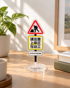 God Is at Work acrylic desktop sign inspired by a road works warning sign, displayed on a wooden desk beside plants and books