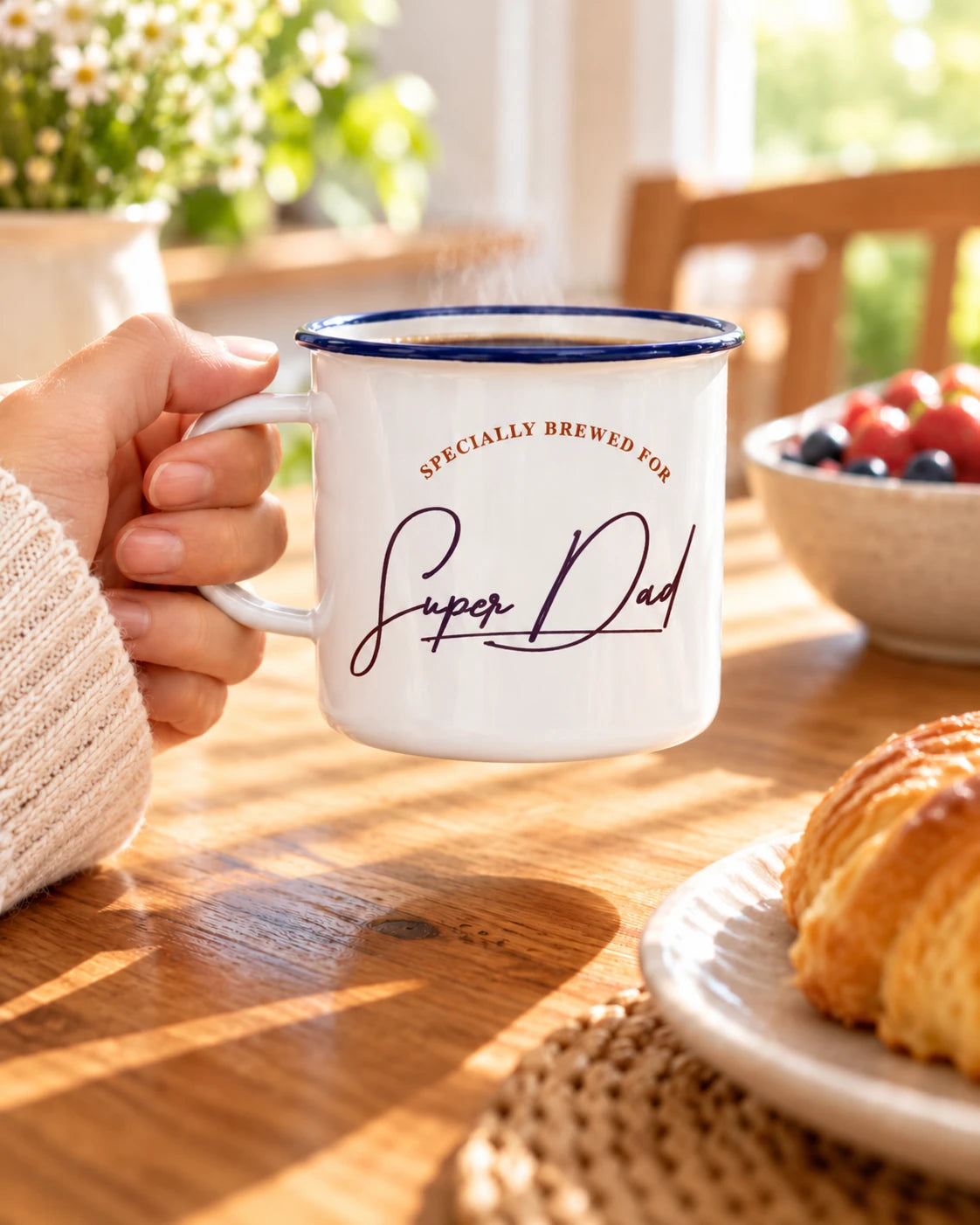 White Super Dad enamel mug with navy rim held by a hand over a cosy breakfast table with pastries and fruit