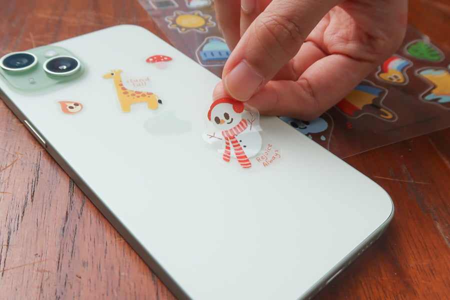 Hand placing a cute weather themed Christian sticker from the everyday believer sticker sheet onto a phone, featuring playful faith-inspired illustrations