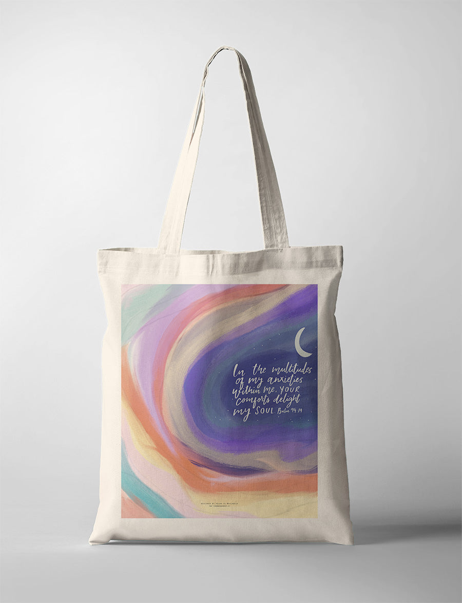 A pastel-colored tote bag showcasing swirling shades of blue, purple, orange, and pink. The design includes a crescent moon and the Bible verse Psalm 94:19: 'In the multitude of my anxieties within me, Your comforts delight my soul.' A modern Christian gift blending practicality and inspiration.