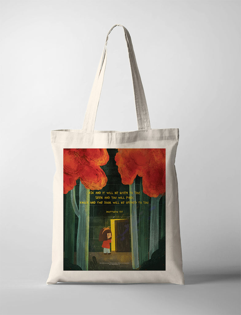 A beige tote bag featuring a Christian-themed illustration of a child standing in front of an open door in a forest, with red trees above. The design includes the Bible verse from Matthew 7:7: 'Ask and it will be given to you; seek and you will find; knock and the door will be opened to you,' printed on the bag, symbolizing faith and guidance.