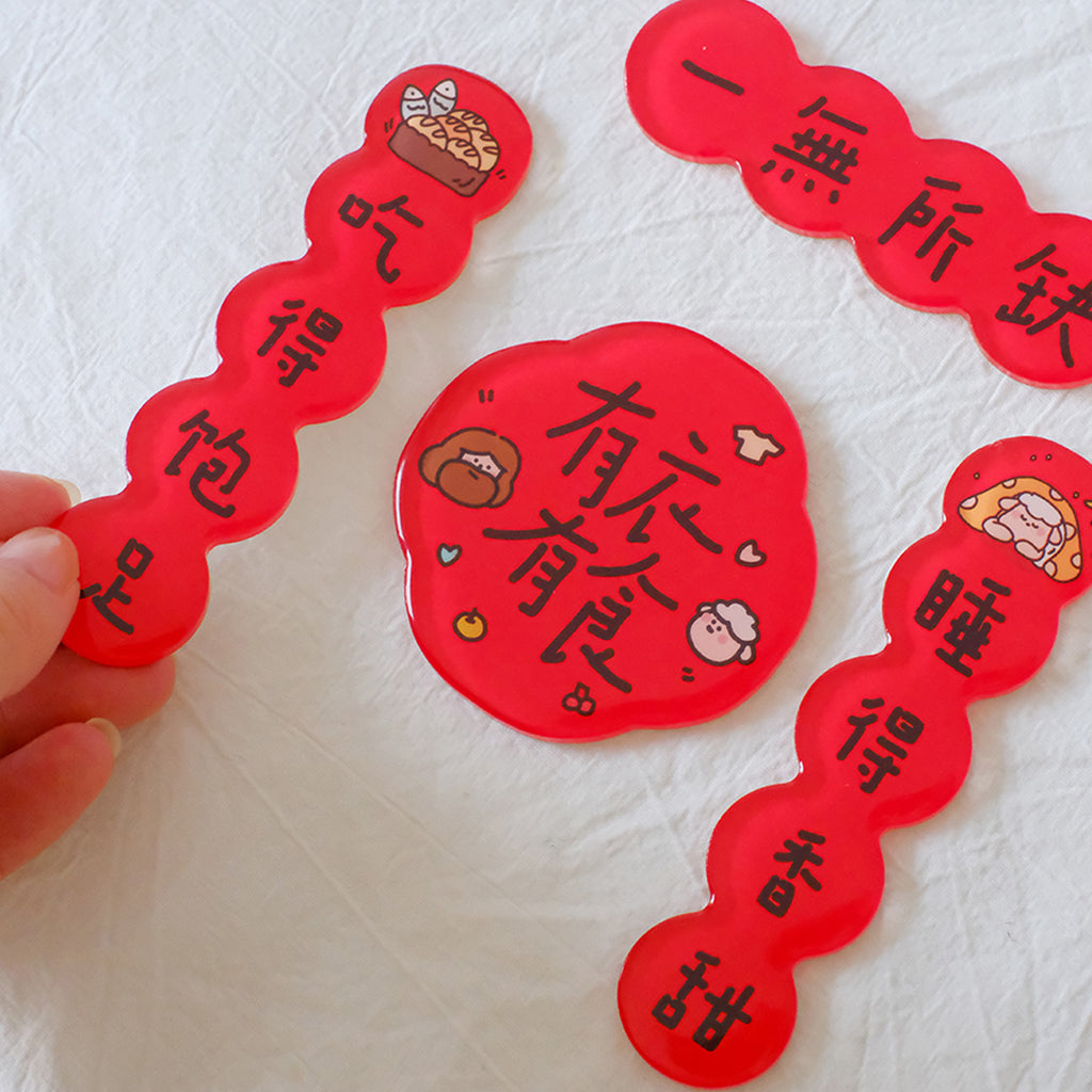 Hand holding a red Chinese New Year couplet magnet with festive blessings, surrounded by matching decorative magnets on a fabric surface