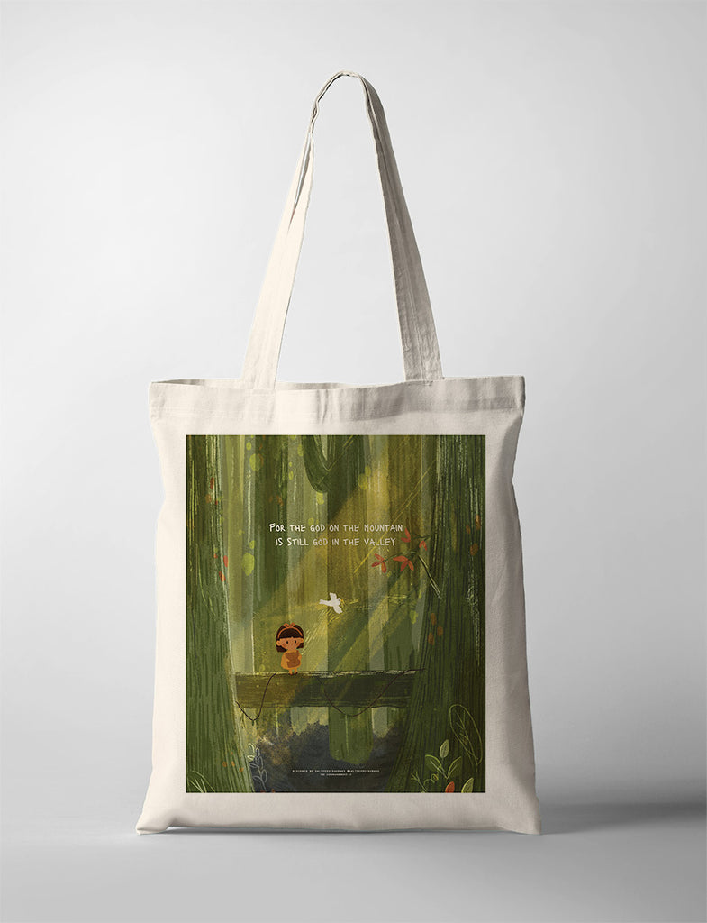 A beige tote bag featuring a Christian-themed illustration of a child standing on a bridge in a forest, with a white bird flying nearby. The design includes the text, 'For the God on the mountain is still God in the valley,' conveying a message of faith and God's presence in all circumstances.