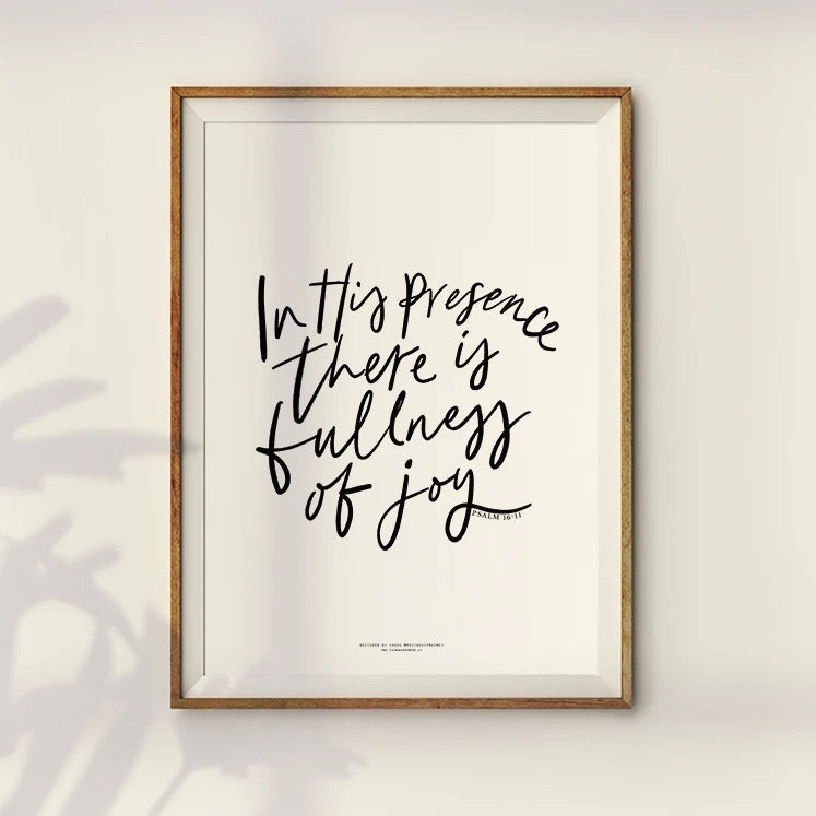 Minimalistic wall art featuring a Bible verse, 'In His presence there is fullness of joy,' written in black calligraphy on a white canvas, surrounded by a wooden frame.