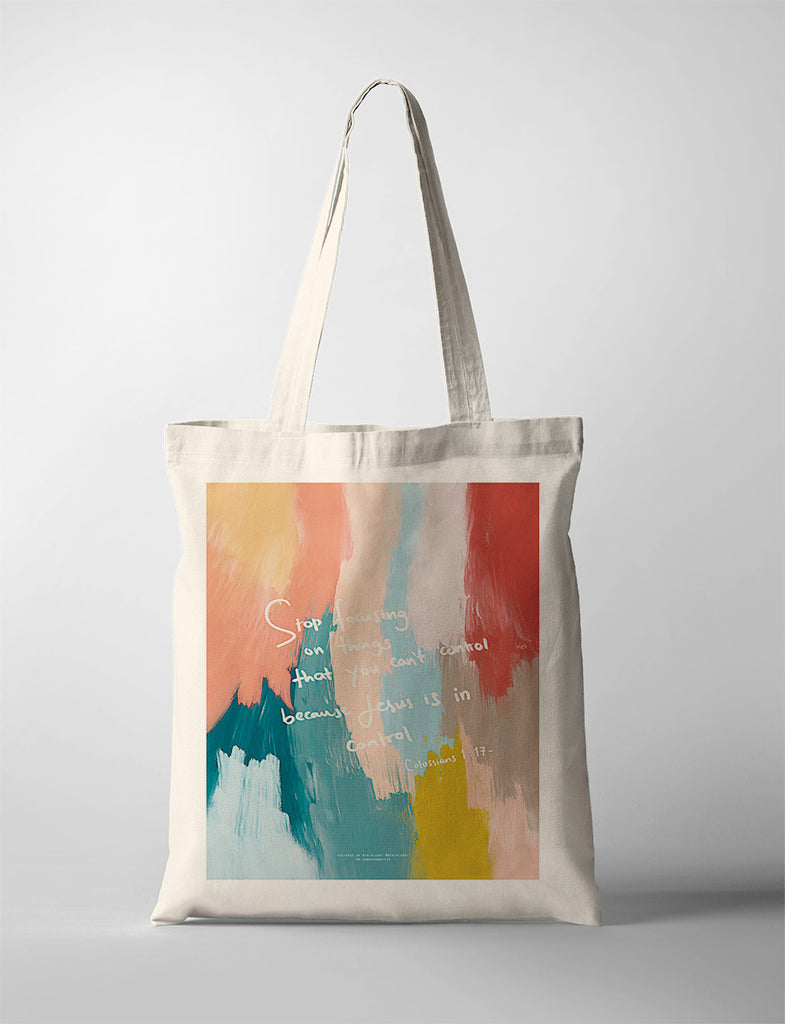 A beige tote bag featuring the empowering quote "Stop focusing on things that you can't control because Jesus is in control" from Colossians 1:17, overlaid on a vibrant and abstract brushstroke design in warm and cool tones.