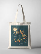 A faith based Tote Bag featuring an inspirational quote, "Led by His Light" displayed on a canvas tote bag. The design includes a glowing lantern amidst a dark teal background, emphasizing themes of hope and guidance. The uplifting message on the faith based poster encourages inner light and positivity