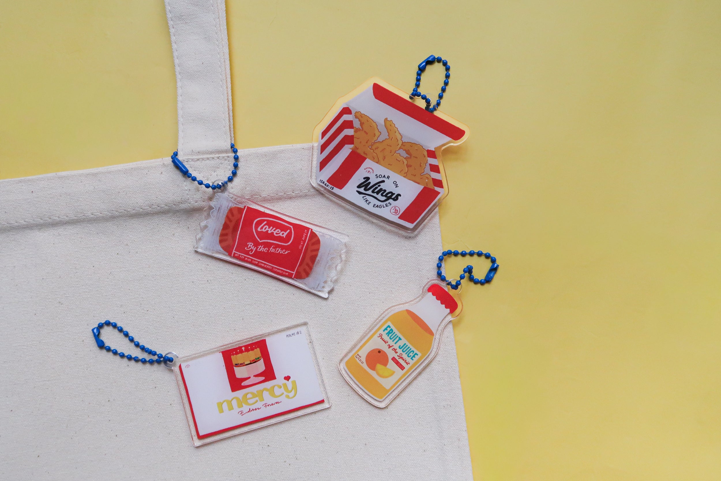Assorted Love Supermarket acrylic keychains attached to a canvas tote bag, featuring designs such as fried chicken wings, loved biscuit, mercy cake, and fruit juice.