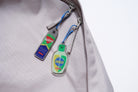 Two Love Supermarket acrylic keychains—BeGone Worries and Armor of God—attached to the zippers of a light grey backpack with blue beaded chains.