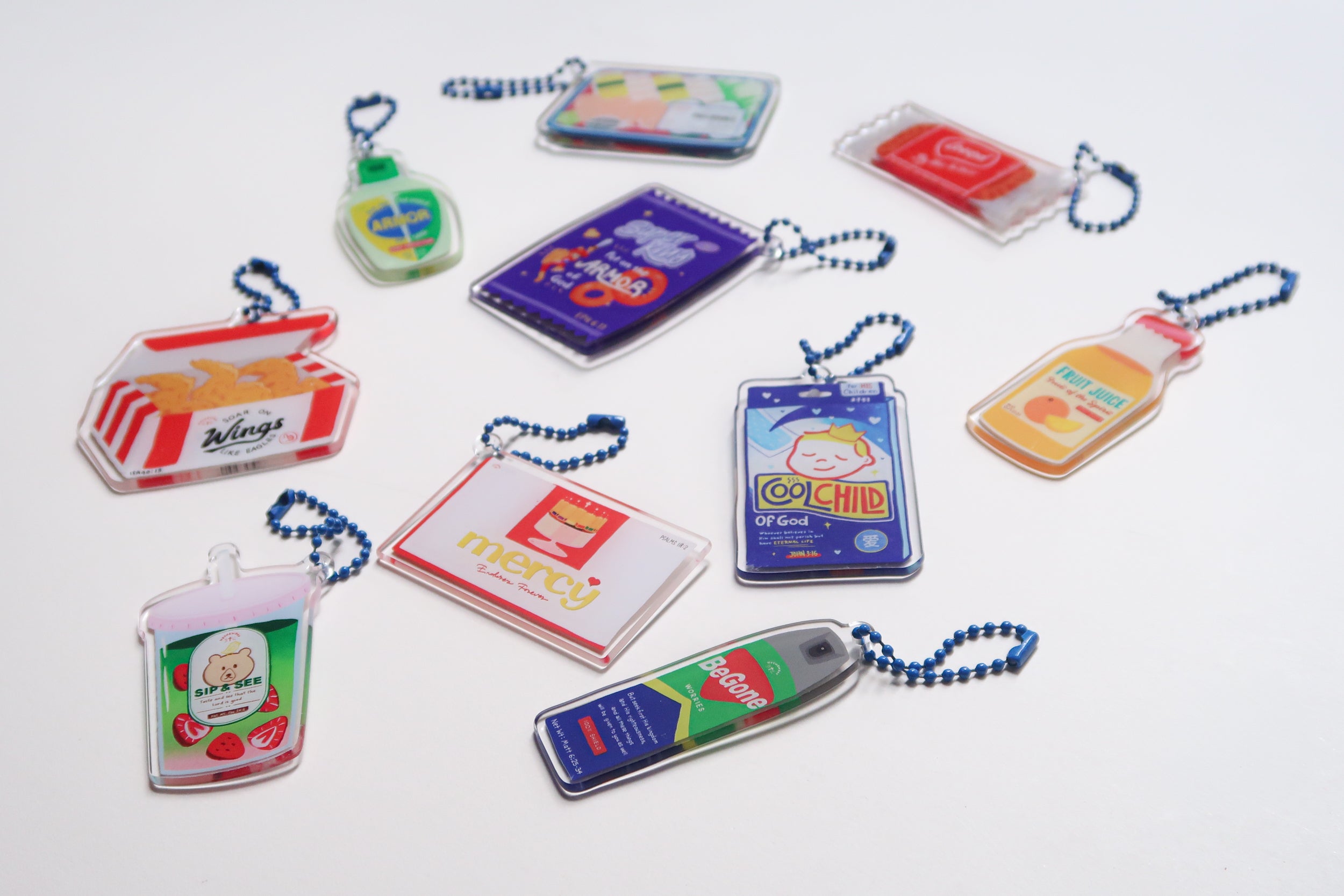 Flat lay of Love Supermarket acrylic keychain collection, including designs like sushi set, Cool Child patch, Armor of God, Begone Worries spray, fried wings, and more.