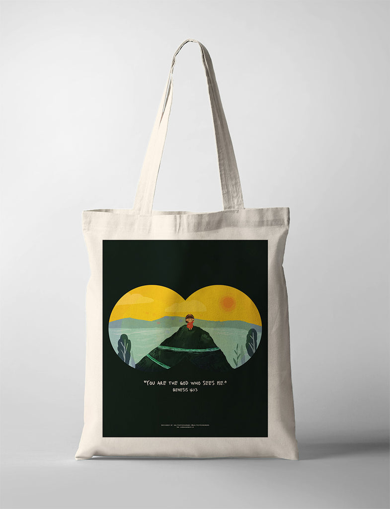 A beige tote bag featuring an illustration of a child sitting on a hilltop with a vibrant yellow and green landscape in the background, symbolized through binoculars. The design includes the Bible verse from Genesis 16:13: 'You are the God who sees me,' conveying a message of being seen and known by God.